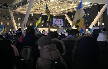 Group of people holdings flags in front of a white, brightly lit stage outdoors.