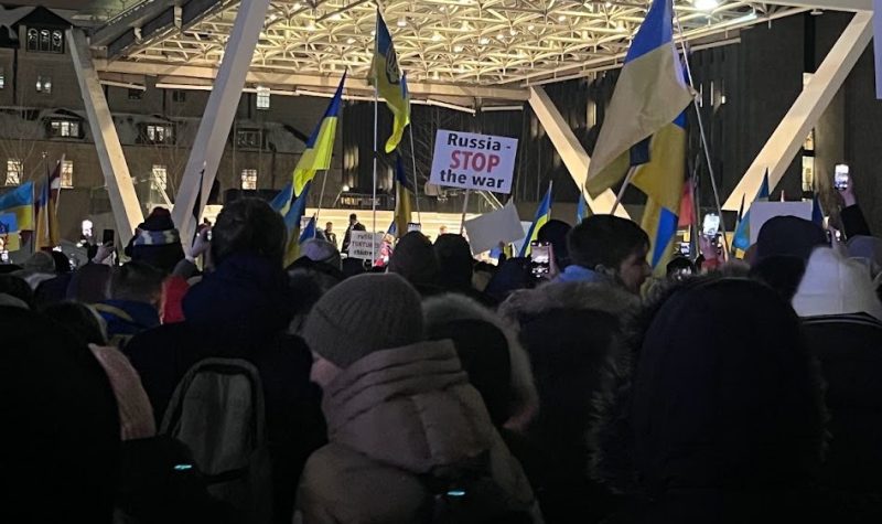 Group of people holdings flags in front of a white, brightly lit stage outdoors.