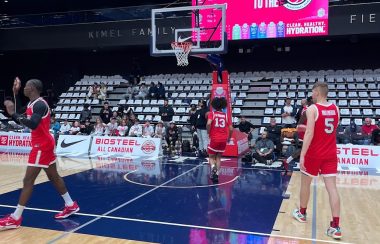 Three people in red jerseys are on a wooden court that is light brown and navy. There is a large red sign on the wall in the background.