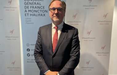 Johan Schitterer stands in front of a white banner at the Bastille Day event. He is wearing a dark grey suit and red tie.