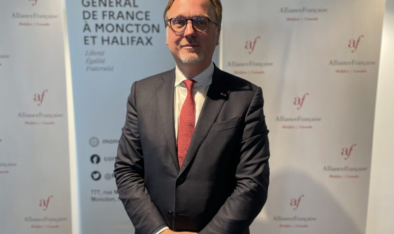 Johan Schitterer stands in front of a white banner at the Bastille Day event. He is wearing a dark grey suit and red tie.