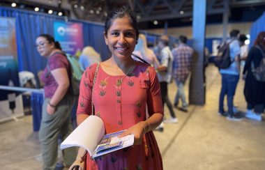 A woman stands in the middle of a job fair holding a booklet. Behind here, there are people and royal blue curtains..