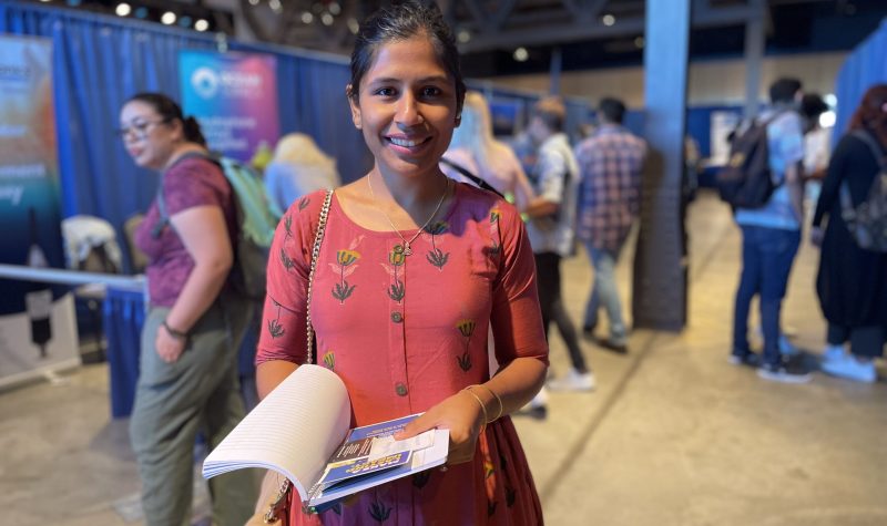 A woman stands in the middle of a job fair holding a booklet. Behind here, there are people and royal blue curtains..