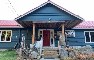 Looking upwards at the front porch and door of a blue one-story building with a red roof.