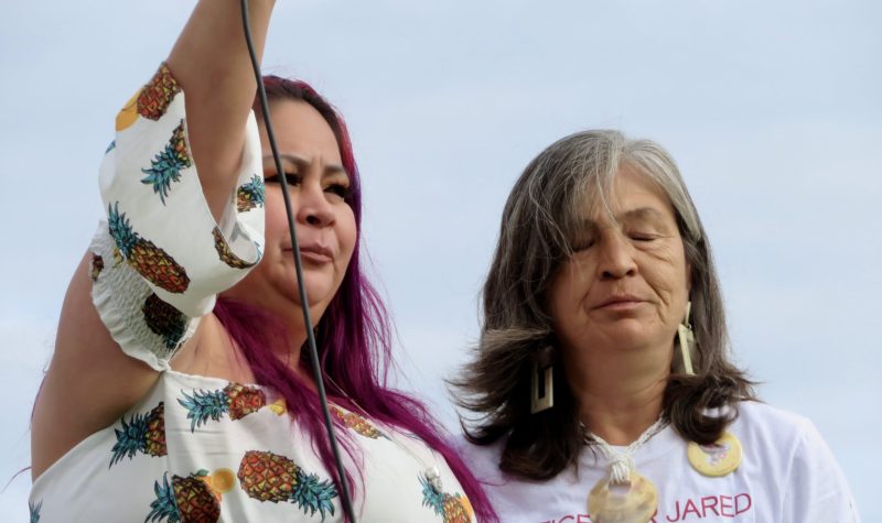 Two Indigenous woman stand together outside at a rally on an overcast day. One of them holds a microphone in the air.