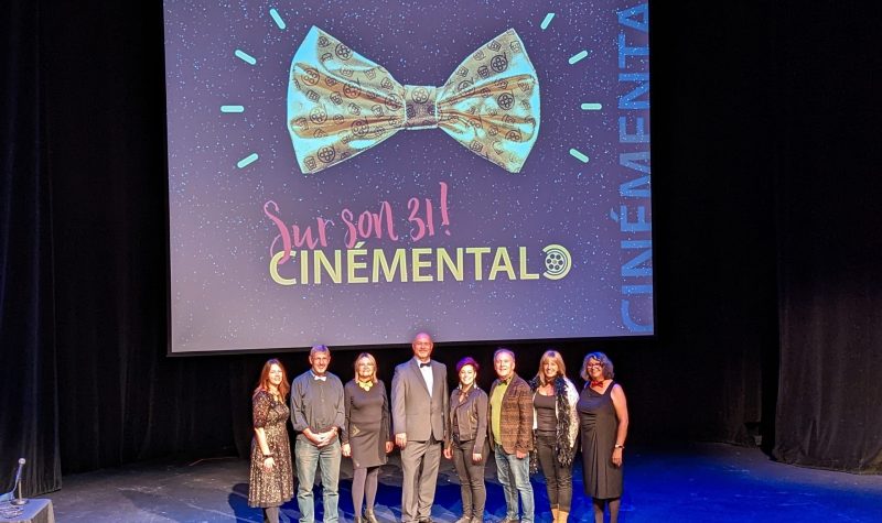 Le président de l'organisation Cinémental, Henri Dupuis, debout au milieu avec de nombreuses autres personnes autour de lui posant devant un grand écran qui indique qu'il s'agit du 31ème festival de films.