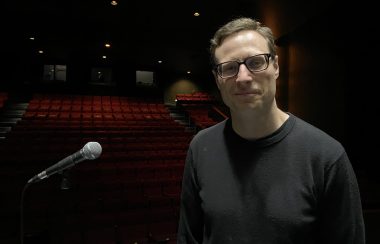 Un homme pose sur scène après un spectacle, à côté d'un micro, avec la salle en arrière plan.