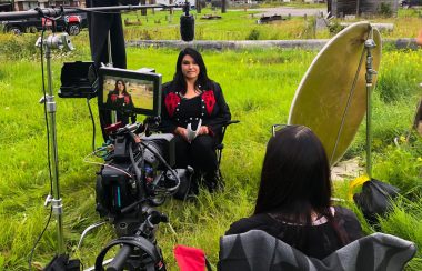 A woman sits in a field on a movie set with camera equipment and other filmmakers surrounding her.