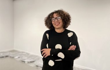 A woman with glasses smiling infront of a blank white wall.