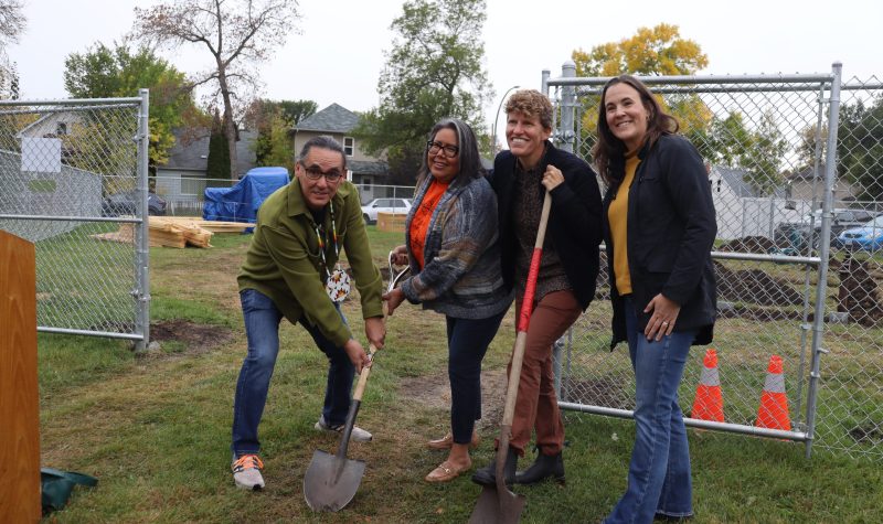 A man and three women stand together on a field. There are orange pilons behind them and the group holds two shovels to the ground. Weather is cloudy.