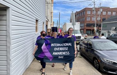 Men and women dressed in purple marching down a street holding a banner that says International Overdose Awareness Day. There are cars and buildings in the background.