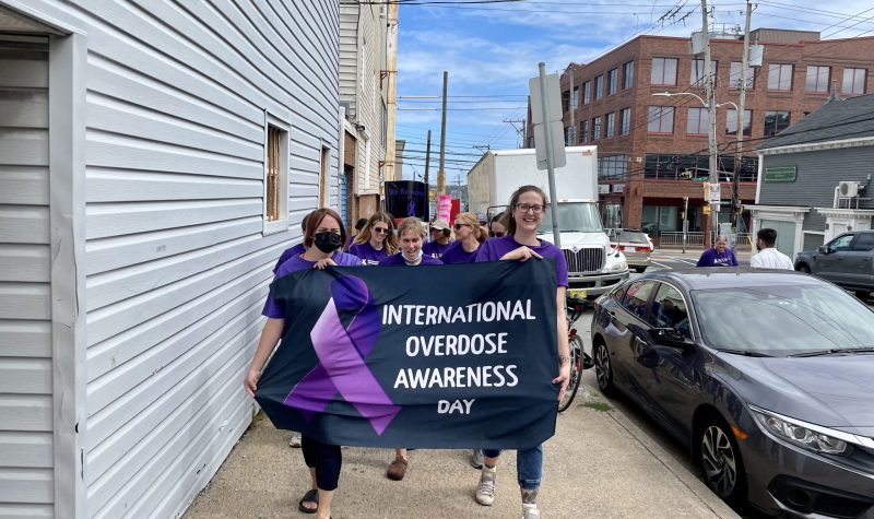 Men and women dressed in purple marching down a street holding a banner that says International Overdose Awareness Day. There are cars and buildings in the background.
