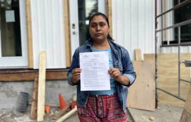 A woman is holding up a paper, there is construction work in the background on her house. She is wearing a jean jacket with her hair tied.