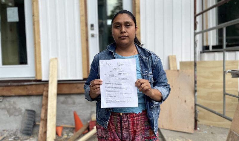 A woman is holding up a paper, there is construction work in the background on her house. She is wearing a jean jacket with her hair tied.