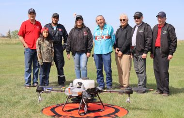 A group of people from the Papaschase First Nation and Papaschase Security Services stand behind a large drone while it sits idle on a small helipad. Weather is clear.
