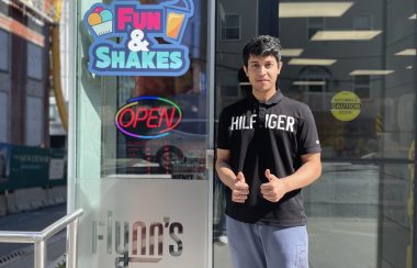 A man in a black shirt standing outside in front of Flynn's Dairy Bar and Convenience. He is holding to thumbs up as he smiles for the camera. There is a sign that says open in red on the store.