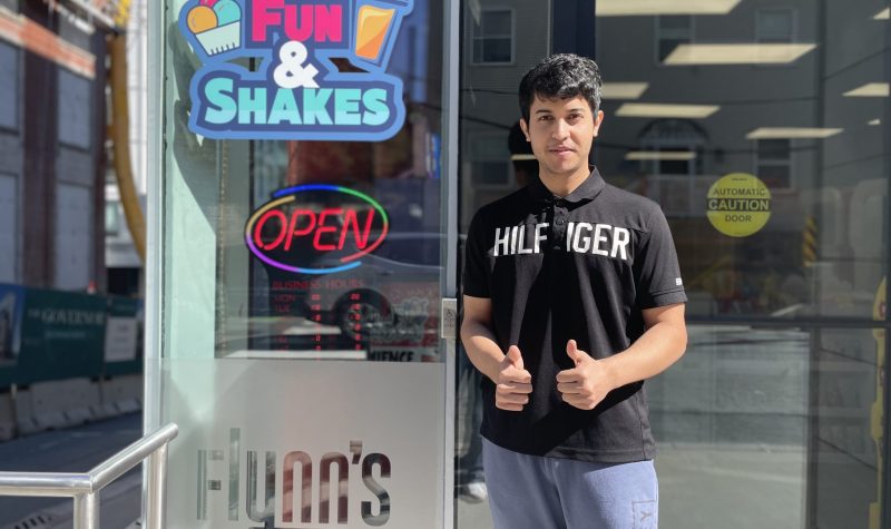 A man in a black shirt standing outside in front of Flynn's Dairy Bar and Convenience. He is holding to thumbs up as he smiles for the camera. There is a sign that says open in red on the store.