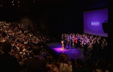 A long shot of a large group of people on stage in an auditorium and audience members facing them. The room is dark. A purple screen can be seen in the background with the Bishop's University logo in white..