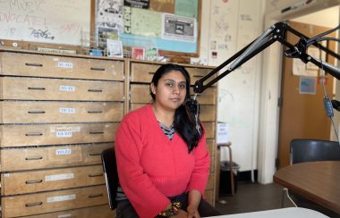 A woman is sitting near a microphone at a studio and looking at the camera, she wearing red and there are drawers behind her.