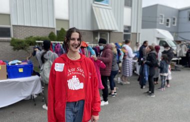 A girl smiling at the camera, she is wearing red and has a name tag with Jacqueline on her. Behind her there are racks of coats, and shoes and people sifting through items at the coat drive.