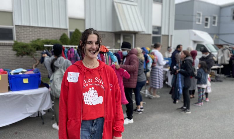 A girl smiling at the camera, she is wearing red and has a name tag with Jacqueline on her. Behind her there are racks of coats, and shoes and people sifting through items at the coat drive.