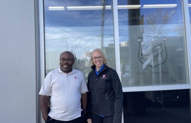 A man and a woman wearing shirts that say Salvation Army on them, they are standing in front of The Salvation Army on Gottingen St. They are both smiling.