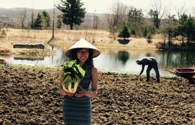 Stéphanie Wang standing in one of the fields at Le Rizen holding an organic Asian vegetable in hand. Behind her is a farmer working the field with a wheel barrow to the side.