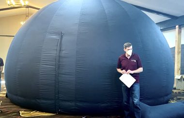 Blue inflatable dome, used as a planetarium with man standing facing camera wearing blue jeans and purple shirt, inside the hall of another building