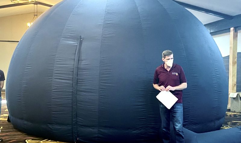 Blue inflatable dome, used as a planetarium with man standing facing camera wearing blue jeans and purple shirt, inside the hall of another building