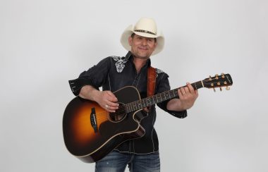 Un homme de style country portant une chemise noir, des jeans bleus et un chapeau blanc tenant sa guitare de couleur noire dégradée