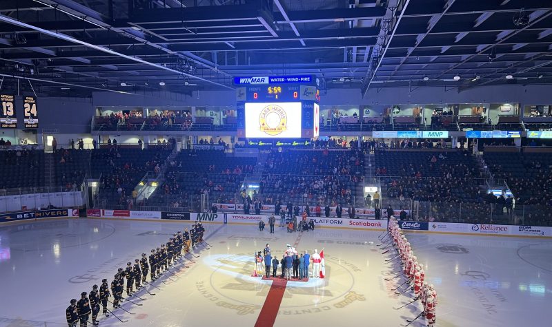 A hockey stadium and rink with Queen's Gaels mens hockey team on the left, RMC Paladins on the right and in between them, prominent figures of the Kingston, Queen's and RMC communities.