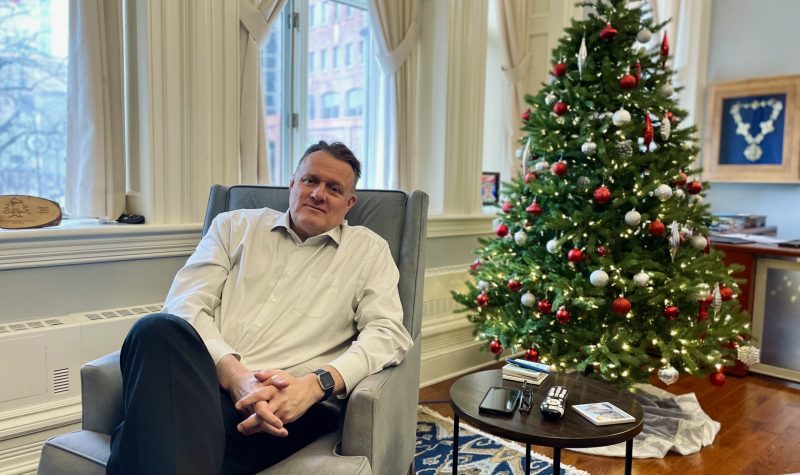 Portrait photo of Halifax Mayor Mike Savage sitting in his office at city hall. There is a christmas tree in the background and he is seen smiling with hands and his legs crossed.