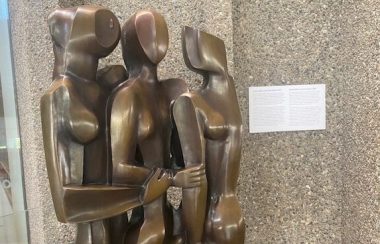 Une statue de bronze représentant trois femmes enlacées, devant un mur gris