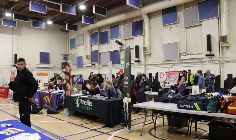 A gymnasium full of tables of agencies offering their services to people in need. There's people walking around, talking to those behind the tables.