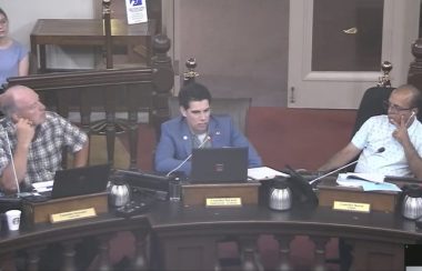 three councillors sit in a row behind the rounded wooden desks in city hall.