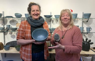 Two women pose with their art pieces in front of shelves of pottery and stone art.