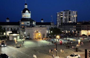 a high-up view of City Hall in Kingston, Ontario at night. Downtown is busy as many people walk in the area surrounding the building.