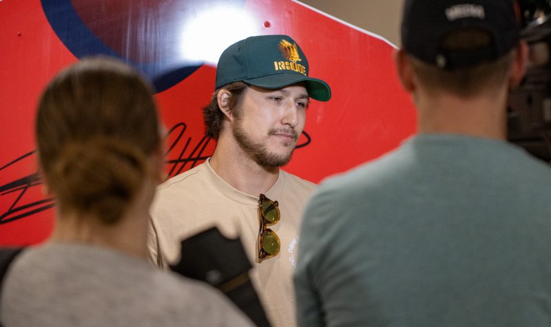 A man in a baseball cap answers questions while looking at two people standing in front of him. He is standing in front of a brightly lit red backdrop with a glaring spotlight reflecting off the backdrop.
