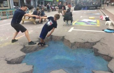 Two people engage with a 3D optical illusion chalk drawing that makes it appear that there is a cracked hole in the road.