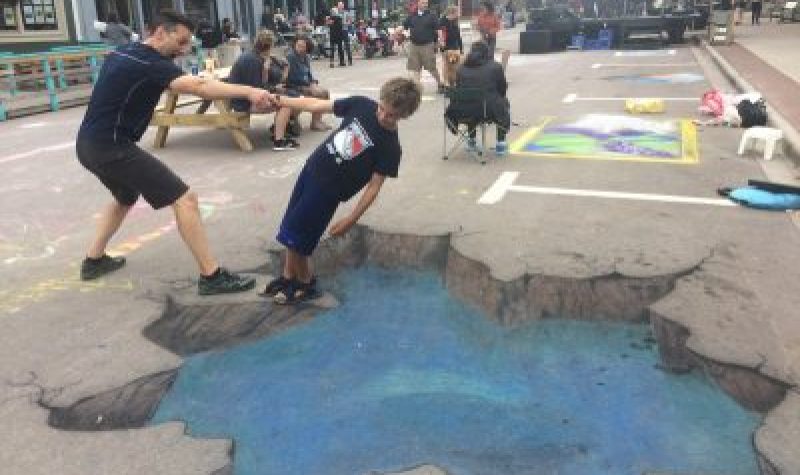 Two people engage with a 3D optical illusion chalk drawing that makes it appear that there is a cracked hole in the road.