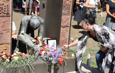 A lady leans over to place a flower on the Homeless Memorial statue in downtown Edmonton during a memorial service. There are a lot of flowers already placed by the statue in all sorts of different colors. Weather is clear.