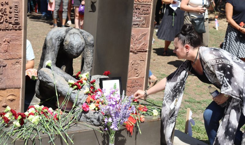 A lady leans over to place a flower on the Homeless Memorial statue in downtown Edmonton during a memorial service. There are a lot of flowers already placed by the statue in all sorts of different colors. Weather is clear.