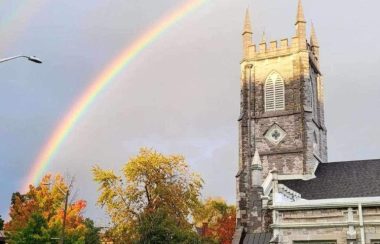 a rainbow in the blue sky over one of the grey stone queen's university buildings. the trees in the bottom left of the frame are a mix of orange and green.