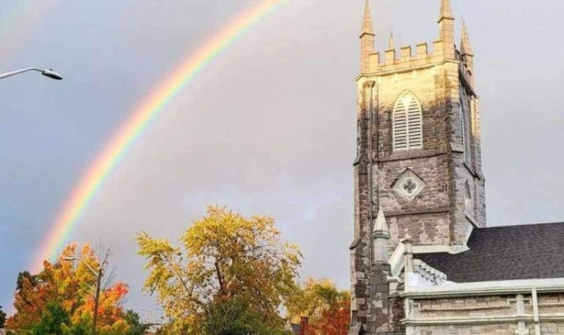 a rainbow in the blue sky over one of the grey stone queen's university buildings. the trees in the bottom left of the frame are a mix of orange and green.