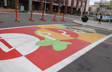 A colorful crosswalk on Okîsikow (Angel) Way in Edmonton, showing Indigenous language and arts. Weather is cloudy.