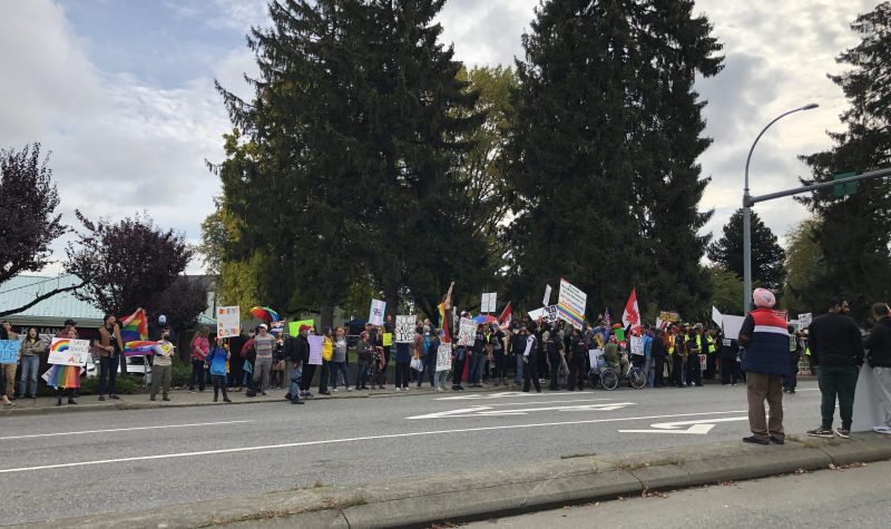 Two groups of people stand side by side on a busy street. On the left the counter-protestors hold signs with rainbows and messages in support of 2SIALGBTQ+ rights. On the right the protestors hold signs demanding SOGI to be removed from education programs