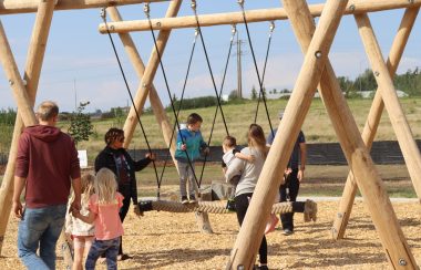 Children playing on a brand new play structure made of wood, with a big piece of string tethered to the top of the structure for kids to walk across and balance themselves on. Weather is clear.