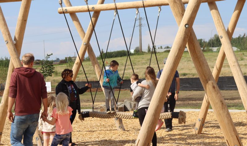 Children playing on a brand new play structure made of wood, with a big piece of string tethered to the top of the structure for kids to walk across and balance themselves on. Weather is clear.