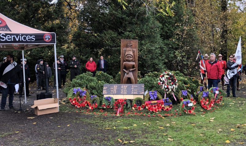 The Sto:lo Nation's plaque and carving stands in a garden of headges. Wreaths decorated with poppies and ribbons for Remembrance Day are planted in front. Members of the Sto:Lo stand on either side, one Elder holds the eagle staff to lead the procession. A forest is in the background.