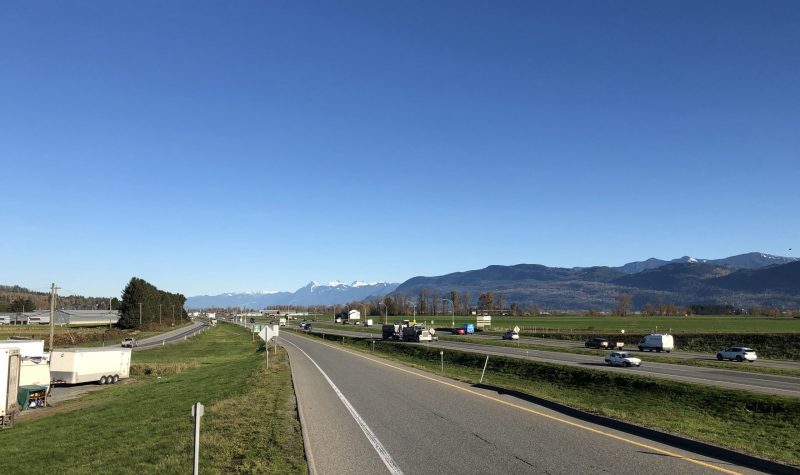 A well travelled highway road exit lined by fields of green grass. Cars and trucks drive on the highway underneath a bright blue sky. Mountains loom in the background wrapping around the highway.